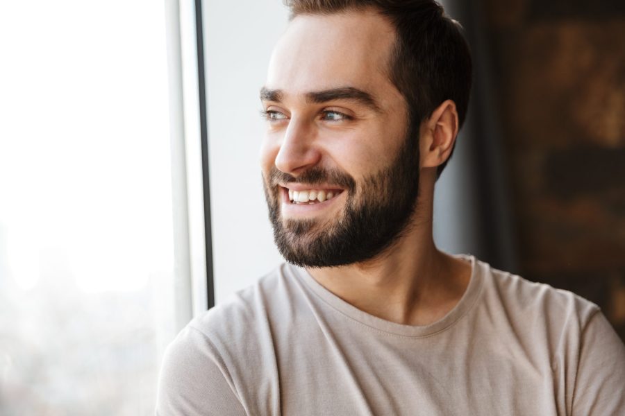 Handsome smiling young bearded man standing at the window at home, looking away